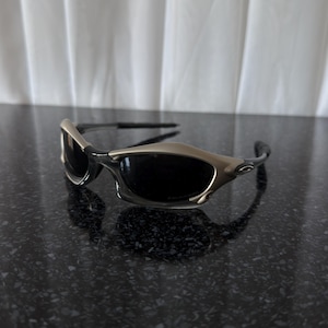 May include: Sport sunglasses with a silver frame and dark lenses. The sunglasses have a sleek, modern design with a curved shape. The lenses are dark, providing protection from the sun. The frame is a metallic silver color.