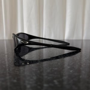 K&ouml;nnte beinhalten: Schwarze Sonnenbrille mit einem schlanken, modernen Design. Die Sonnenbrille hat einen gebogenen Rahmen und dunkle Gl&auml;ser. Das Oakley-Logo ist an der Seite des Rahmens sichtbar. Sie liegt auf einer dunklen, reflektierenden Oberfl&auml;che.