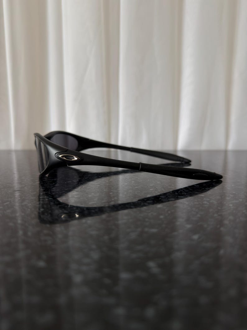 K&ouml;nnte beinhalten: Schwarze Sonnenbrille mit einem schlanken, modernen Design. Die Sonnenbrille hat einen gebogenen Rahmen und dunkle Gl&auml;ser. Das Oakley-Logo ist an der Seite des Rahmens sichtbar. Sie liegt auf einer dunklen, reflektierenden Oberfl&auml;che.