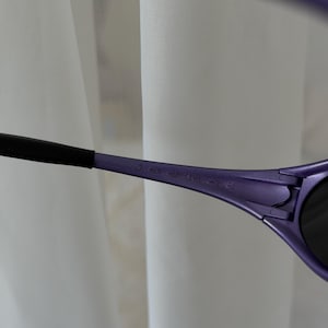 May include: Close-up of purple sunglasses with black arms. The text "jaguar" is printed on the arm of the sunglasses. The sunglasses have dark lenses and a sleek, modern design.