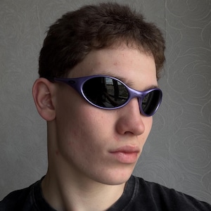 Op de afbeelding: Paarse zonnebril met donkere glazen. De zonnebril heeft een strak, gebogen ontwerp. De afbeelding toont de zonnebril op een persoon, met de focus op de bril.