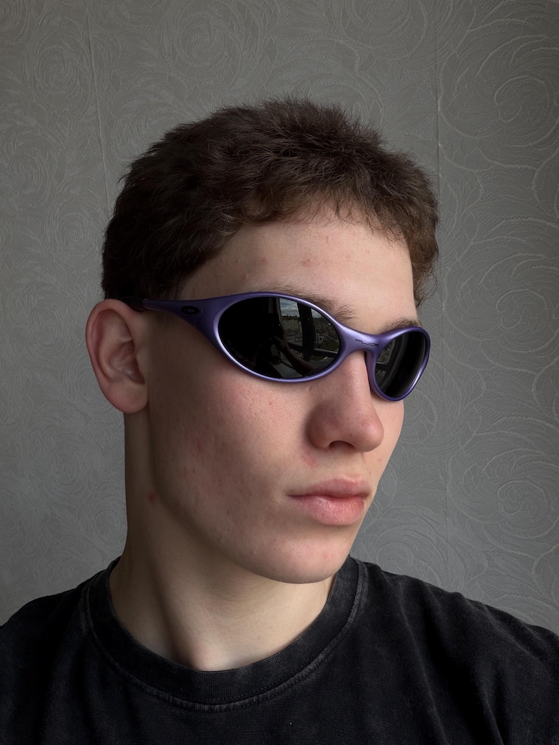 May include: Purple-framed sunglasses with dark lenses. The sunglasses have a sleek, curved design. The image shows the sunglasses on a person, with the focus on the eyewear.