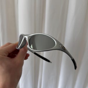 May include: Silver-framed sunglasses with gray lenses. The sunglasses have a curved design and black accents on the temples. The lenses are oval-shaped and reflect the light. The frame has a metallic finish.