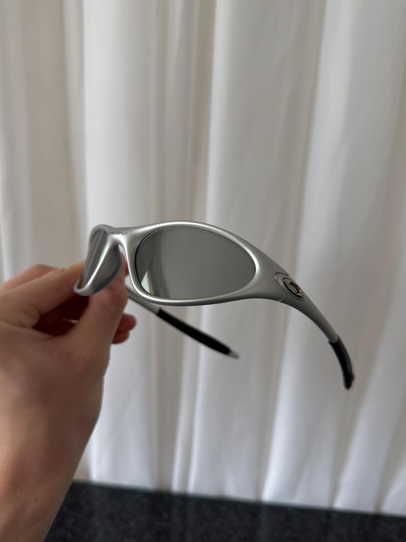 May include: Silver-framed sunglasses with gray lenses. The sunglasses have a curved design and black accents on the temples. The lenses are oval-shaped and reflect the light. The frame has a metallic finish.