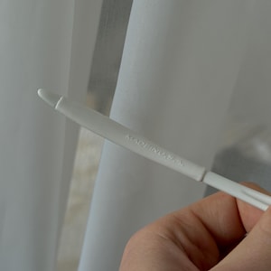 Puede incluir: Primer plano de cortinas blancas transparentes con un gancho de cortina de pl&aacute;stico blanco. El texto "MADE IN U.S.A." est&aacute; impreso en el gancho.