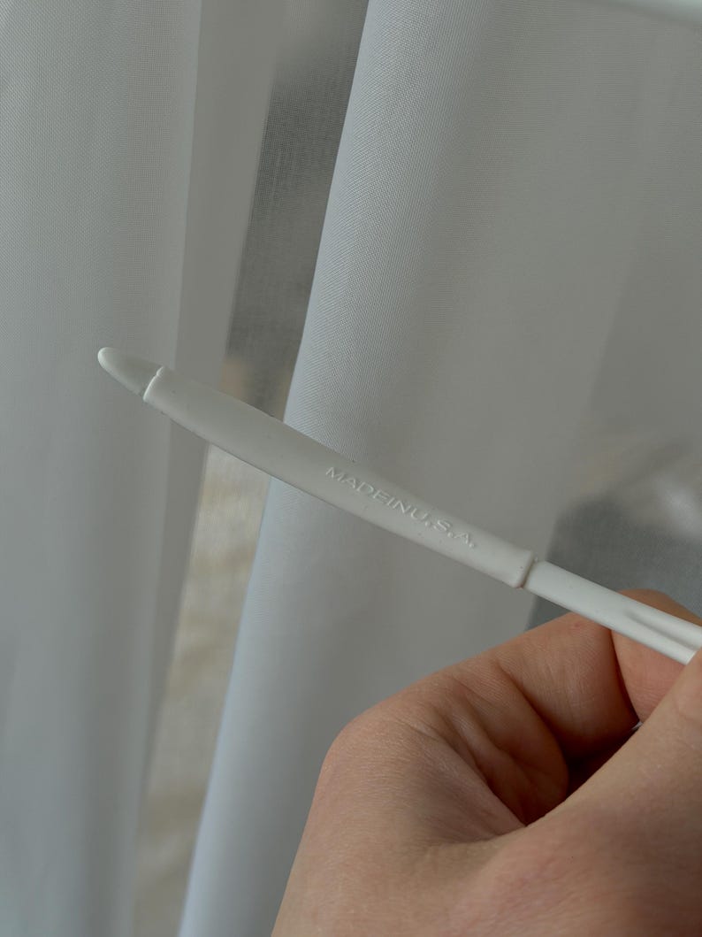 Puede incluir: Primer plano de cortinas blancas transparentes con un gancho de cortina de pl&aacute;stico blanco. El texto "MADE IN U.S.A." est&aacute; impreso en el gancho.