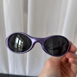 May include: Purple oval sunglasses with dark lenses. The sunglasses have a glossy finish and the word "GOLF" is printed on the bridge. The sunglasses are held in a hand.
