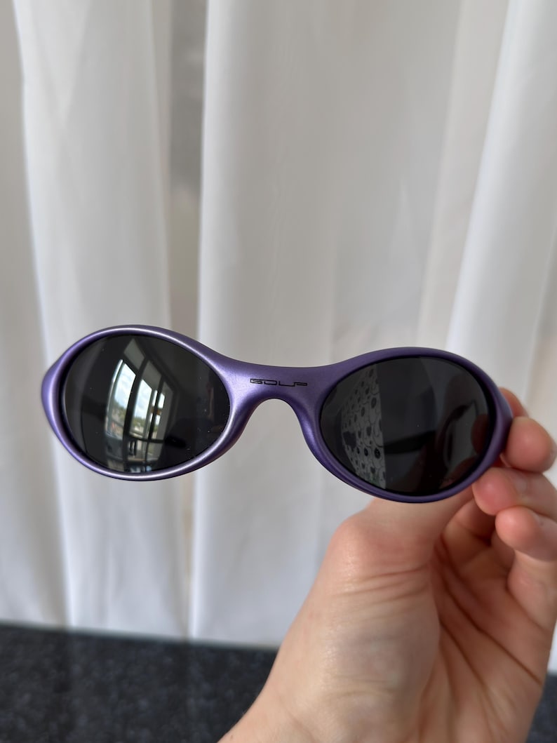 May include: Purple oval sunglasses with dark lenses. The sunglasses have a glossy finish and the word "GOLF" is printed on the bridge. The sunglasses are held in a hand.