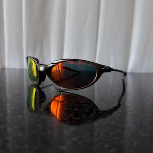 May include: Black sunglasses with a curved frame and reflective lenses. The lenses have a gradient of colors, including yellow, orange, and red. The sunglasses are resting on a dark, reflective surface.