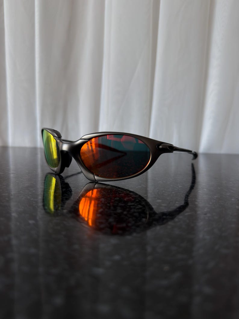 May include: Black sunglasses with a curved frame and reflective lenses. The lenses have a gradient of colors, including yellow, orange, and red. The sunglasses are resting on a dark, reflective surface.