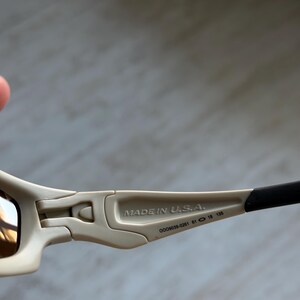 May include: Close-up of sunglasses with a beige frame and dark lenses. The text "MADE IN U.S.A." is printed on the arm. The arm also has the numbers "009039-0261 61 18 135".