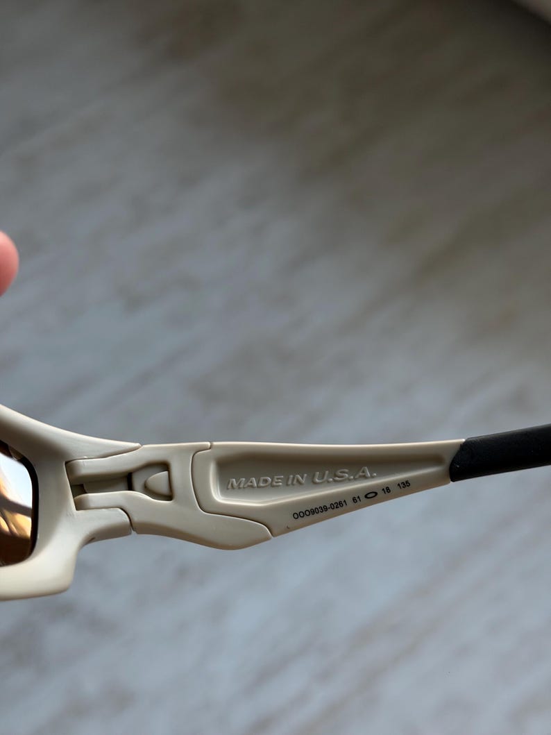 May include: Close-up of sunglasses with a beige frame and dark lenses. The text "MADE IN U.S.A." is printed on the arm. The arm also has the numbers "009039-0261 61 18 135".