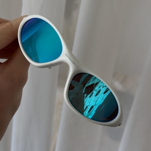 Puede incluir: Gafas de sol blancas con lentes de espejo azules. Las gafas de sol tienen un dise&ntilde;o de montura curvada. Las lentes reflejan un color azul y turquesa. Sobre un fondo blanco.