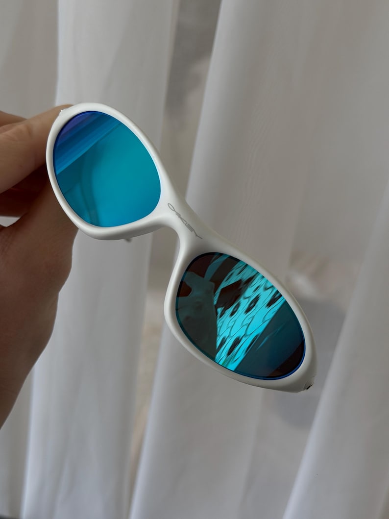 Puede incluir: Gafas de sol blancas con lentes de espejo azules. Las gafas de sol tienen un dise&ntilde;o de montura curvada. Las lentes reflejan un color azul y turquesa. Sobre un fondo blanco.
