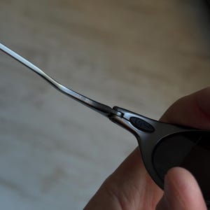 May include: Close-up of a pair of sunglasses with a dark gray frame and dark lenses. The metal arms are thin and sleek, with the number "04 102" printed on the arm. The hinge features a small, stylized logo.