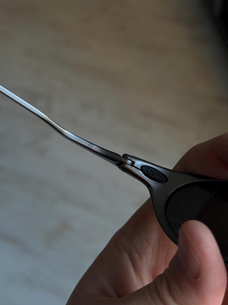 May include: Close-up of a pair of sunglasses with a dark gray frame and dark lenses. The metal arms are thin and sleek, with the number "04 102" printed on the arm. The hinge features a small, stylized logo.