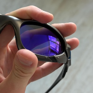 Op de afbeelding: Close-up van een zonnebril met een donkergrijs montuur en blauwe glazen. De glazen reflecteren een raam. De zonnebril heeft een strak, modern design met een metalen montuur en donkergrijze pootjes.