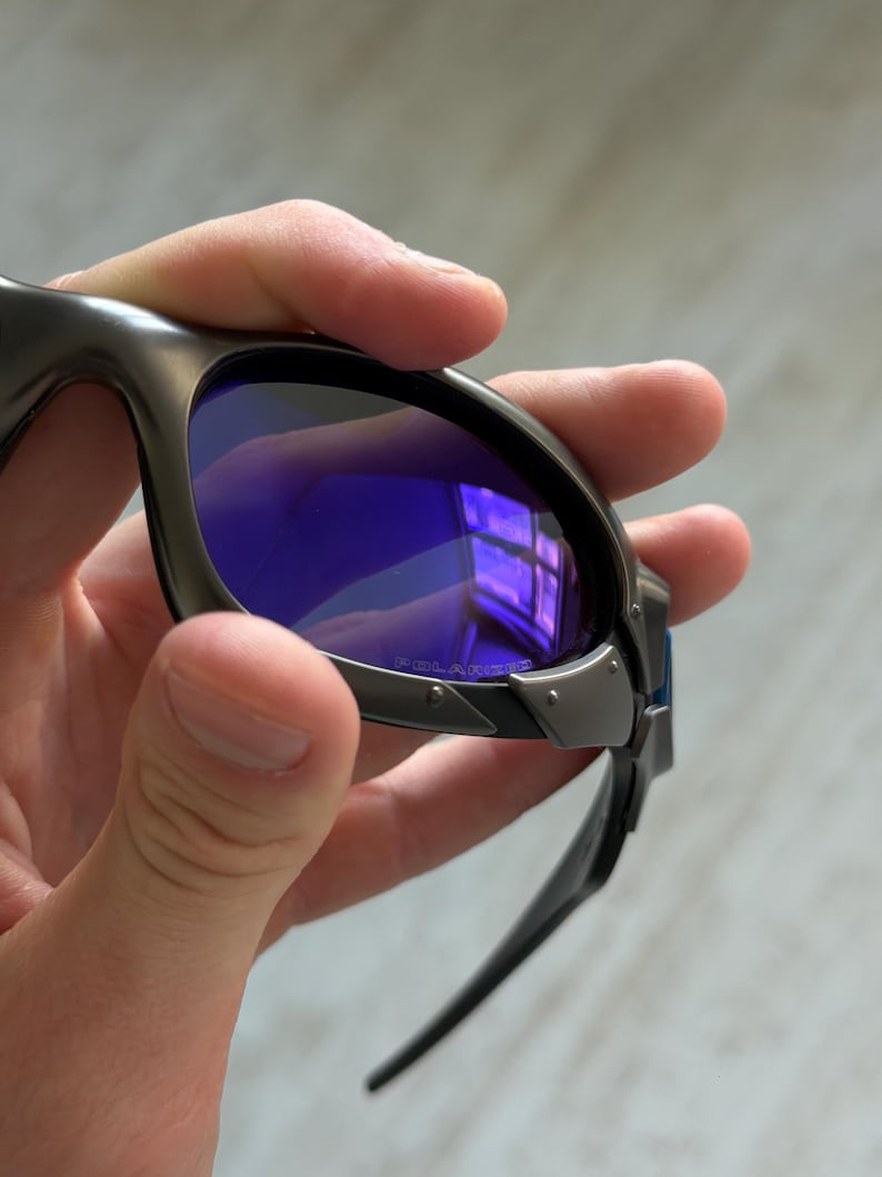 Op de afbeelding: Close-up van een zonnebril met een donkergrijs montuur en blauwe glazen. De glazen reflecteren een raam. De zonnebril heeft een strak, modern design met een metalen montuur en donkergrijze pootjes.