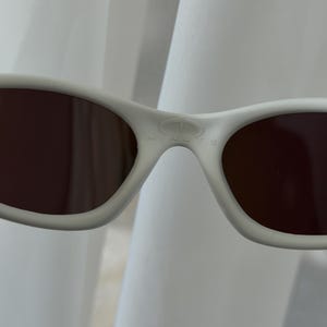Puede incluir: Primer plano de gafas de sol blancas con lentes marrones. La palabra "MINUTE" est&aacute; impresa en el puente de las gafas de sol. Las gafas de sol tienen un dise&ntilde;o curvo y est&aacute;n sobre un fondo blanco.