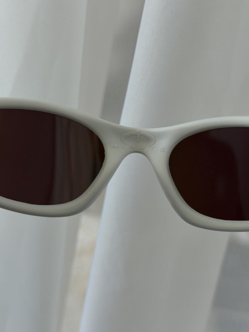 Puede incluir: Primer plano de gafas de sol blancas con lentes marrones. La palabra "MINUTE" est&aacute; impresa en el puente de las gafas de sol. Las gafas de sol tienen un dise&ntilde;o curvo y est&aacute;n sobre un fondo blanco.