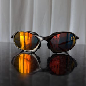 May include: Black sunglasses with orange mirrored lenses. The sunglasses have a sleek, modern design with a dark frame. The lenses reflect a vibrant orange and yellow hue, creating a striking visual effect. The sunglasses are resting on a reflective surface.
