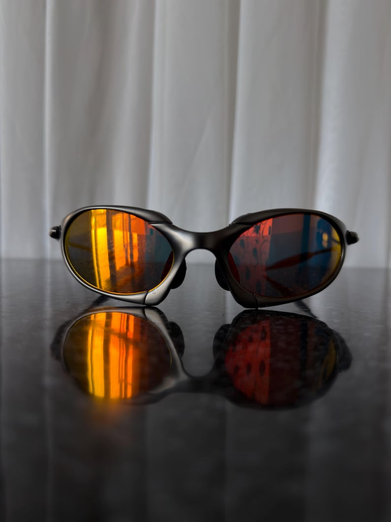 May include: Black sunglasses with orange mirrored lenses. The sunglasses have a sleek, modern design with a dark frame. The lenses reflect a vibrant orange and yellow hue, creating a striking visual effect. The sunglasses are resting on a reflective surface.