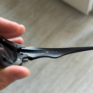 Op de afbeelding: Close-up van een zwarte zonnebril met donkere glazen. De zonnebril heeft een strak, modern design met een gebogen montuur. De tekst "MADE IN U.S.A." is zichtbaar op de poot van de bril.