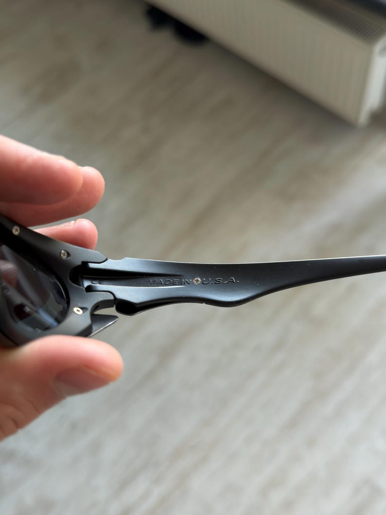 Op de afbeelding: Close-up van een zwarte zonnebril met donkere glazen. De zonnebril heeft een strak, modern design met een gebogen montuur. De tekst "MADE IN U.S.A." is zichtbaar op de poot van de bril.