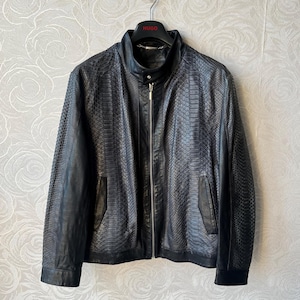 May include: Black leather jacket with a snakeskin pattern, featuring a stand-up collar, a front zipper, and two side pockets. The jacket is hanging on a black hanger with the brand name "HUGO".