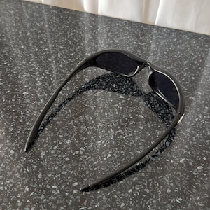 K&ouml;nnte beinhalten: Schwarze Sonnenbrille mit einem schlanken, modernen Design. Die Sonnenbrille hat dunkle Gl&auml;ser und einen geschwungenen Rahmen. Die B&uuml;gel der Sonnenbrille sind d&uuml;nn und reichen bis zu den Schl&auml;fen. Die Sonnenbrille liegt auf einer dunklen, gesprenkelten Oberfl&auml;che.