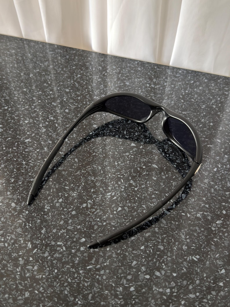 K&ouml;nnte beinhalten: Schwarze Sonnenbrille mit einem schlanken, modernen Design. Die Sonnenbrille hat dunkle Gl&auml;ser und einen geschwungenen Rahmen. Die B&uuml;gel der Sonnenbrille sind d&uuml;nn und reichen bis zu den Schl&auml;fen. Die Sonnenbrille liegt auf einer dunklen, gesprenkelten Oberfl&auml;che.