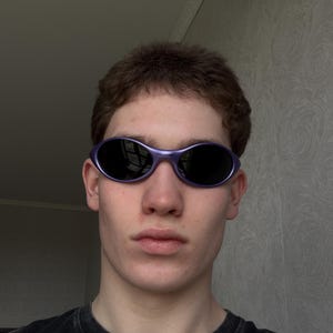 May include: Oval-shaped sunglasses with a purple frame and dark lenses. The sunglasses are worn by a person. The person is wearing a black t-shirt. The background is a light gray wall.