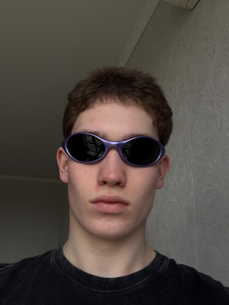 May include: Oval-shaped sunglasses with a purple frame and dark lenses. The sunglasses are worn by a person. The person is wearing a black t-shirt. The background is a light gray wall.
