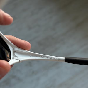May include: White sunglasses with black lenses and arms. The text "CE MADE IN U.S.A." is printed on the arm of the sunglasses. The sunglasses are held by a person.