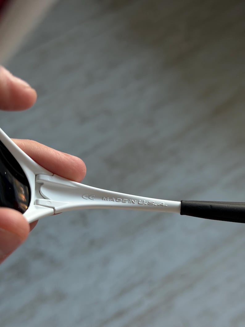 May include: White sunglasses with black lenses and arms. The text "CE MADE IN U.S.A." is printed on the arm of the sunglasses. The sunglasses are held by a person.