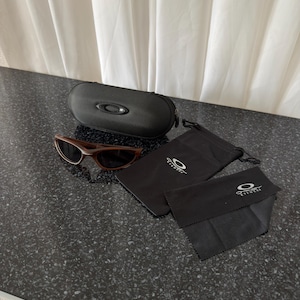 May include: Brown sunglasses with a black case, a black drawstring bag, and a black cleaning cloth. The sunglasses have a dark brown frame and lenses. The case is oval-shaped with a zipper closure. The accessories have the Oakley Eyewear logo.