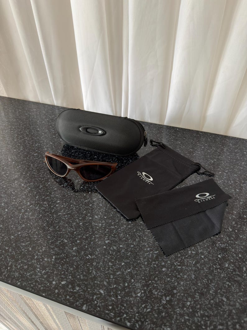 May include: Brown sunglasses with a black case, a black drawstring bag, and a black cleaning cloth. The sunglasses have a dark brown frame and lenses. The case is oval-shaped with a zipper closure. The accessories have the Oakley Eyewear logo.