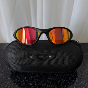 May include: Black sunglasses with red and orange mirrored lenses rest on a black, textured case. The sunglasses have a sleek, sporty design. The case features the brand's logo. The lenses reflect the surrounding light.