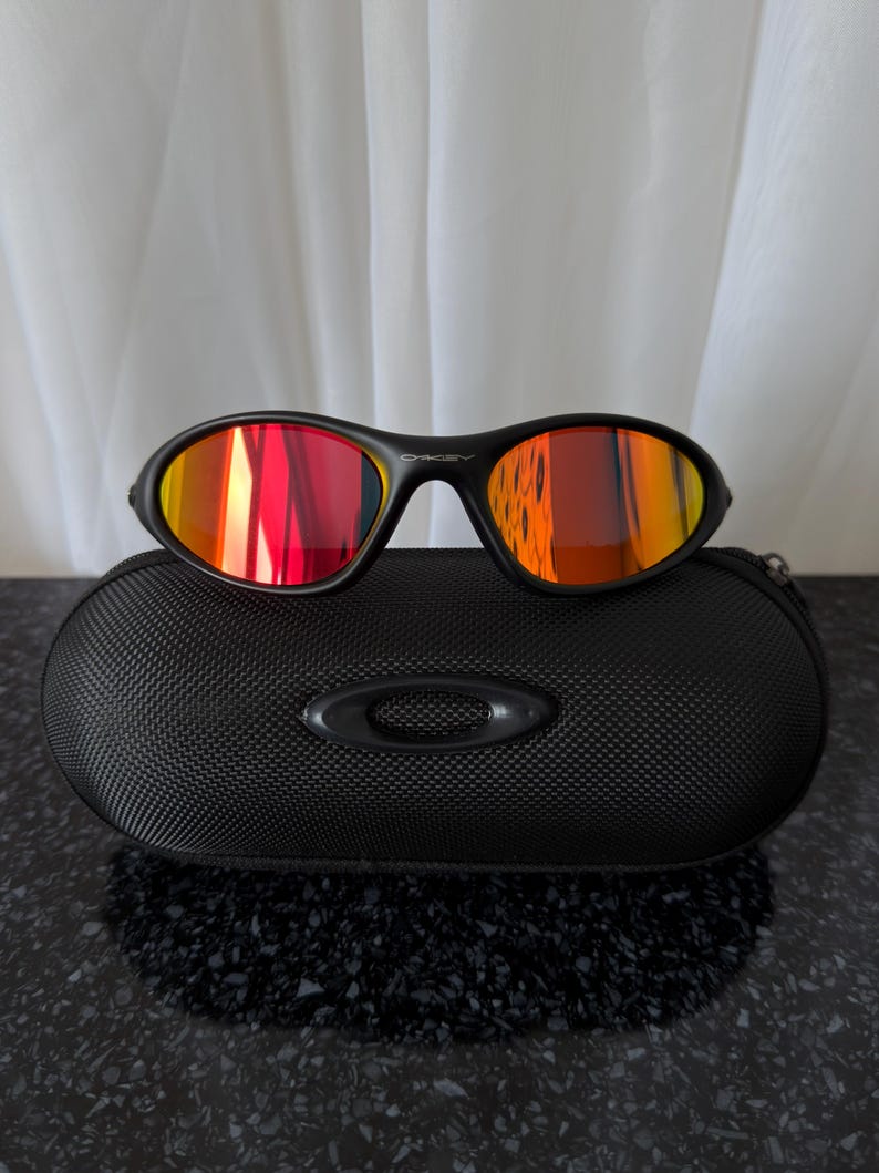 May include: Black sunglasses with red and orange mirrored lenses rest on a black, textured case. The sunglasses have a sleek, sporty design. The case features the brand's logo. The lenses reflect the surrounding light.