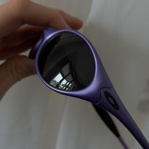 May include: Purple sunglasses with dark lenses. The sunglasses have a sleek, curved design with a matte finish. The lenses reflect the surrounding environment. The brand name is visible on the lens.