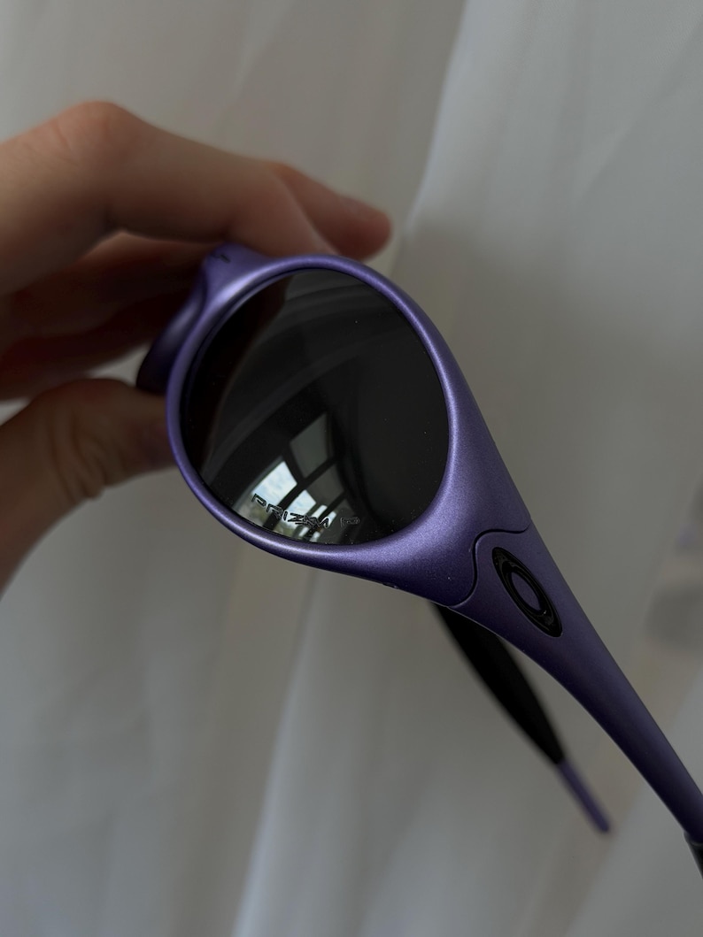 May include: Purple sunglasses with dark lenses. The sunglasses have a sleek, curved design with a matte finish. The lenses reflect the surrounding environment. The brand name is visible on the lens.