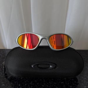 Puede incluir: Gafas de sol con montura plateada y lentes espejadas rojas y amarillas sobre un estuche negro texturizado. Las gafas tienen un dise&ntilde;o elegante y el estuche presenta un logotipo. Las lentes reflejan colores vibrantes.