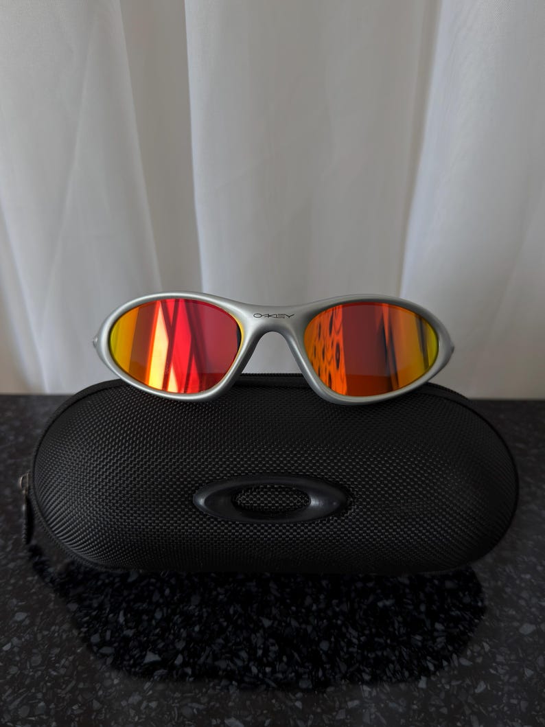 Puede incluir: Gafas de sol con montura plateada y lentes espejadas rojas y amarillas sobre un estuche negro texturizado. Las gafas tienen un dise&ntilde;o elegante y el estuche presenta un logotipo. Las lentes reflejan colores vibrantes.