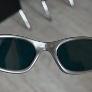 Puede incluir: Primer plano de gafas de sol plateadas con lentes oscuras. La palabra "MINUTE" est&aacute; impresa en el puente. Las gafas de sol tienen un dise&ntilde;o elegante y moderno.