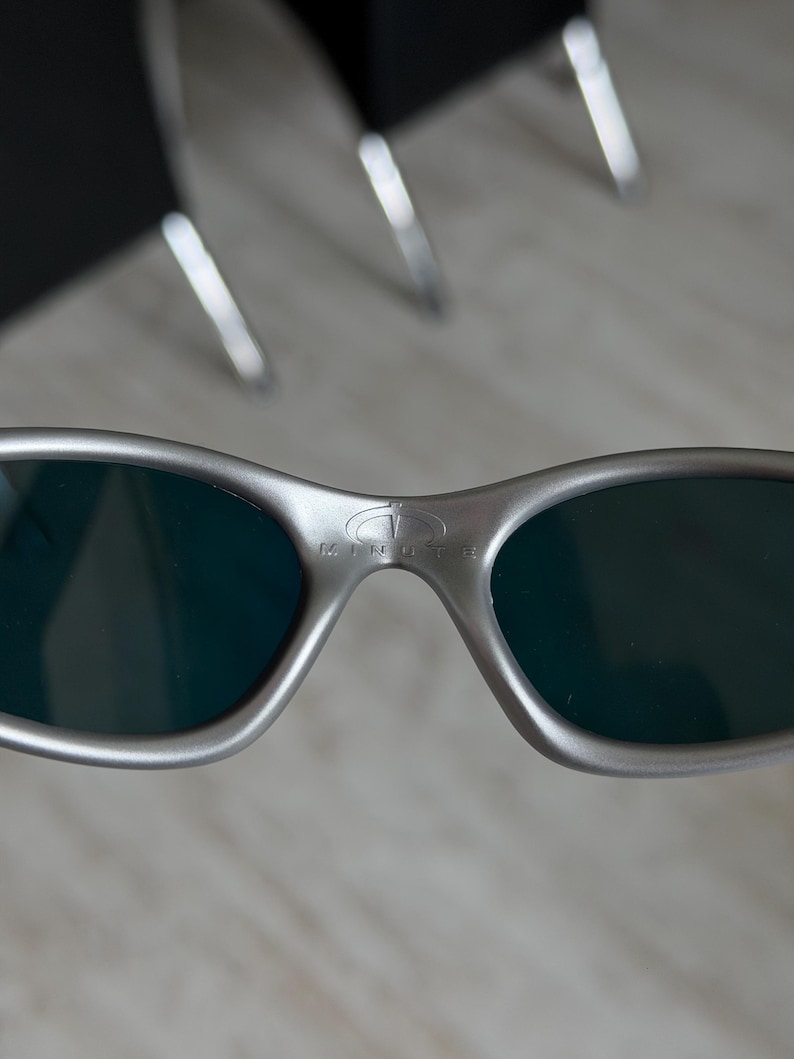 Puede incluir: Primer plano de gafas de sol plateadas con lentes oscuras. La palabra "MINUTE" est&aacute; impresa en el puente. Las gafas de sol tienen un dise&ntilde;o elegante y moderno.