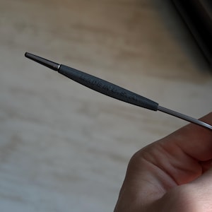May include: A metal knitting needle with a dark gray handle. The handle has the text "MADE IN U.S.A." printed on it. The needle is held in a hand.