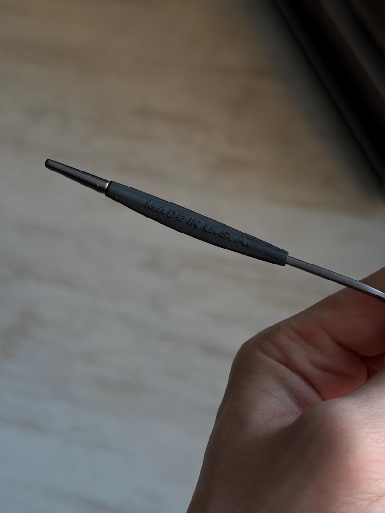 May include: A metal knitting needle with a dark gray handle. The handle has the text "MADE IN U.S.A." printed on it. The needle is held in a hand.
