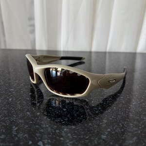 May include: Tan-colored sunglasses with dark lenses. The sunglasses have a curved, sporty design with a thick frame and black arms. The Oakley logo is visible on the side of the frame. The sunglasses are sitting on a dark, reflective surface.