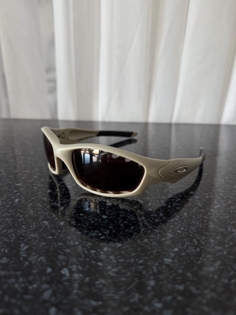 May include: Tan-colored sunglasses with dark lenses. The sunglasses have a curved, sporty design with a thick frame and black arms. The Oakley logo is visible on the side of the frame. The sunglasses are sitting on a dark, reflective surface.