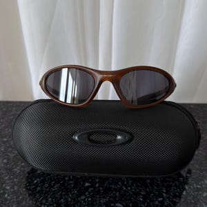 Könnte beinhalten: Braune Sonnenbrille mit dunklen Gläsern liegt auf einem schwarzen, strukturierten Etui. Die Sonnenbrille hat einen geschwungenen Rahmen und der Markenname "Oakley" ist sichtbar. Das Etui hat einen Reißverschluss und ein erhabenes Logo.
